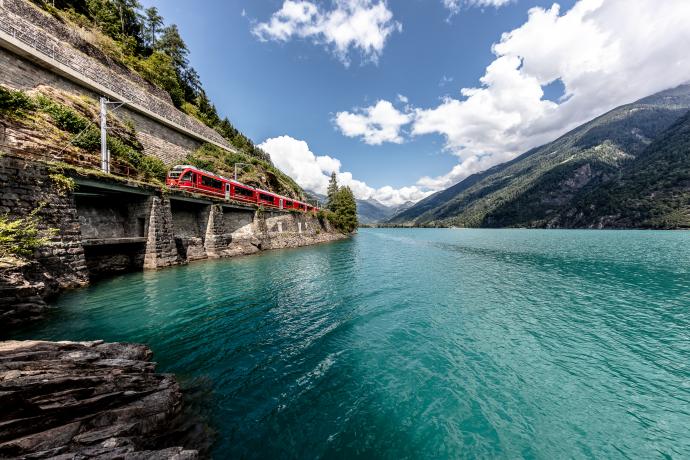 Der Lago di Poschiavo ist ein natürlicher Stausee inmitten des Flusses Poschiavino (Foto: © Rhätische Bahn / Andrea Badrutt) Ein Regionalzug der Rhätischen Bahn fährt am Lago di Poschiavo entlang
